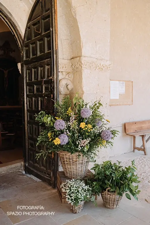 Ambientación floral para bodas en iglesia con cestas de mimbre decoradas con alliums morados, margaritas blancas, solidagos amarillos y follaje verde